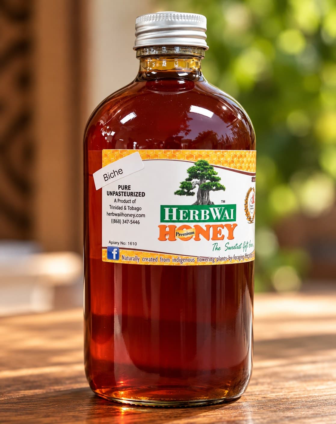 Round bottle of deep red-amber HerbWai Biche pure unpasteurized honey with the bonsai label, on a wooden table