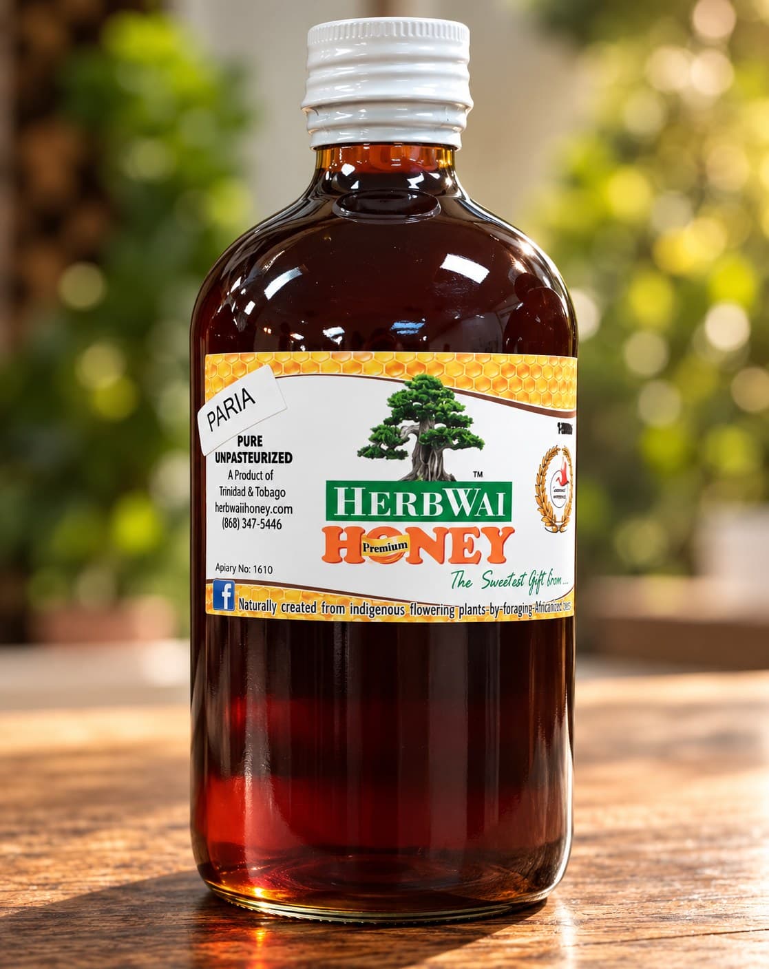 Round bottle of dark amber HerbWai Paria pure unpasteurized honey with the bonsai label, on a wooden table against greenery